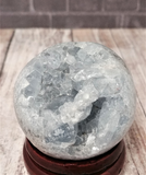 Blue Celestite Sphere on wood stand over wood grain in front of brick Gypsy Gems & Jewelry